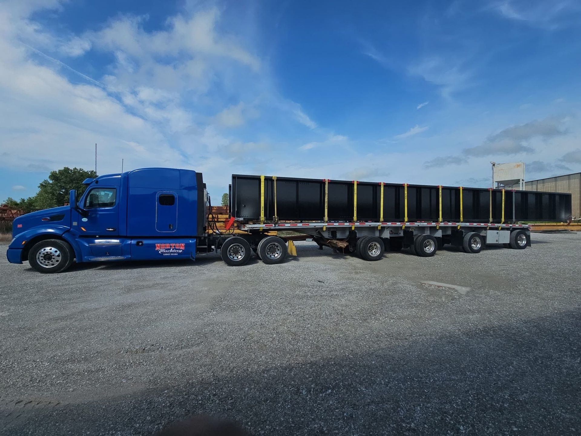 Blue semi-truck hauling multiple black containers on a flatbed trailer against a blue sky.