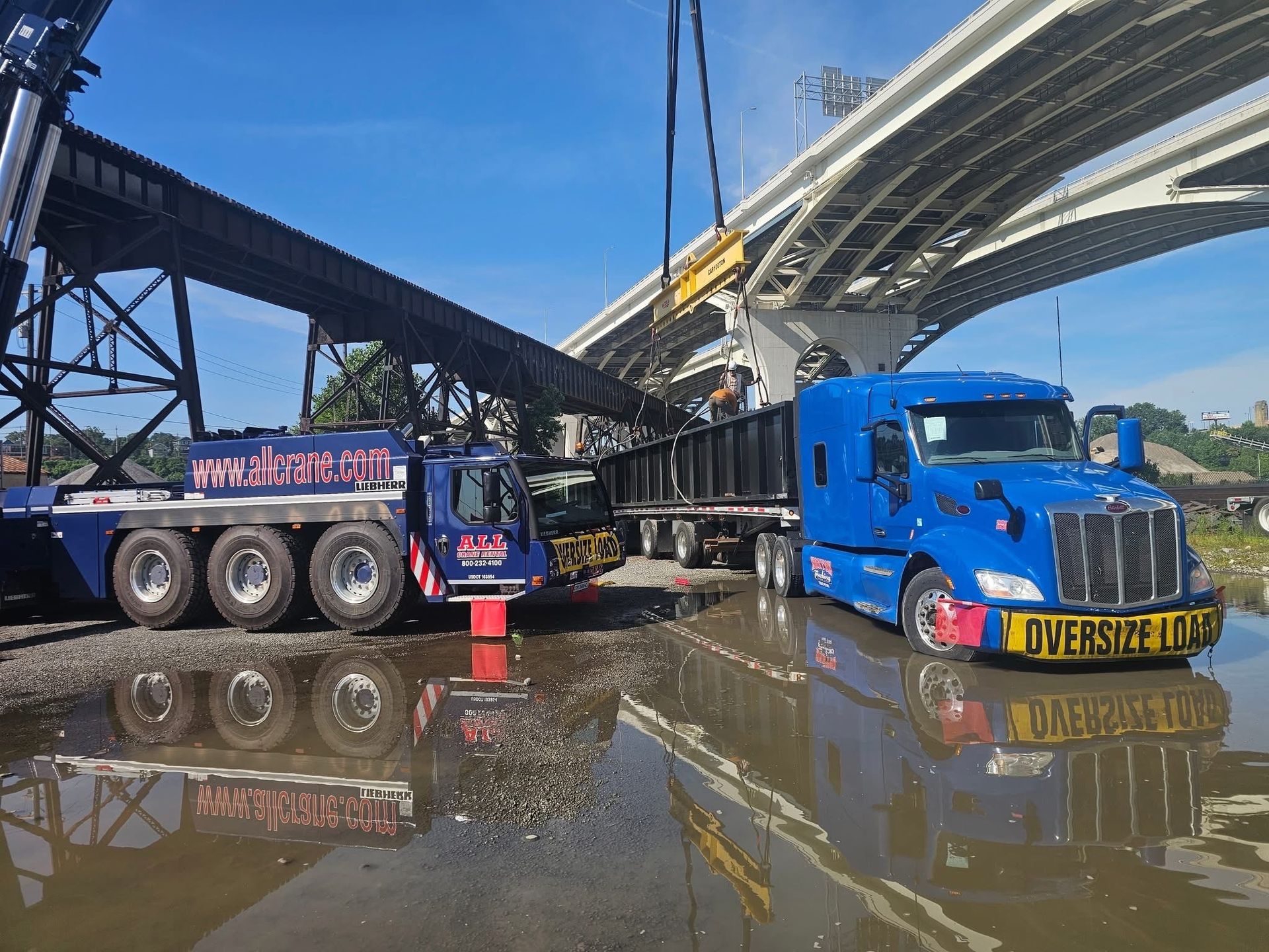 Trucks under a bridge, one hauling a trailer, crane lifting, water on the ground, blue sky.