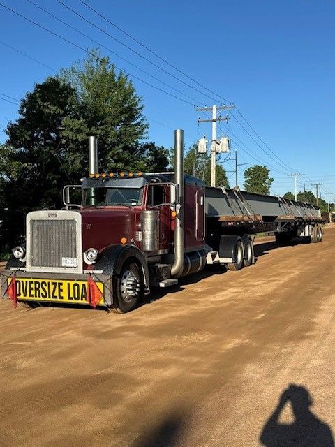 A maroon semi-truck with an oversized load on a flatbed trailer. The truck is parked on a dirt road in a sunny environment.
