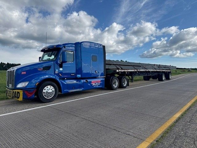 Blue semi-truck with a long flatbed trailer carrying steel beams on a road under a cloudy sky.