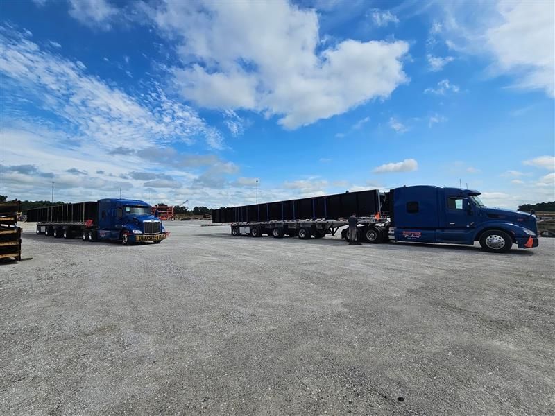 Two blue semi-trucks with flatbed trailers on a gravel lot under a blue, cloudy sky.