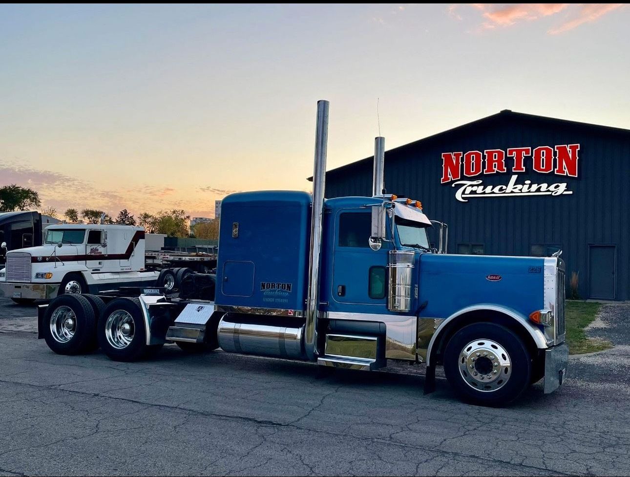 Blue semi-truck parked in front of a building with a red sign; gravel ground. Sunset sky in the background.