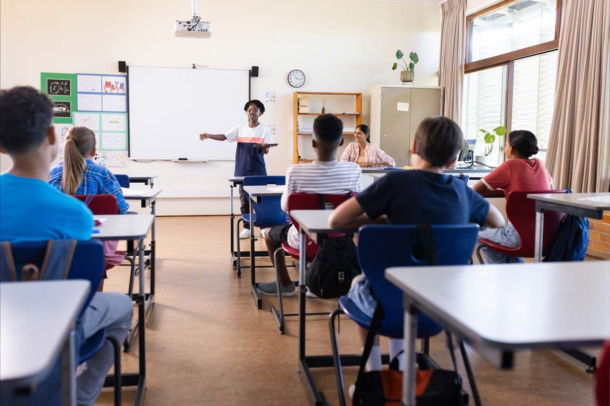 Classroom with students in desks, teacher at whiteboard presenting lesson.