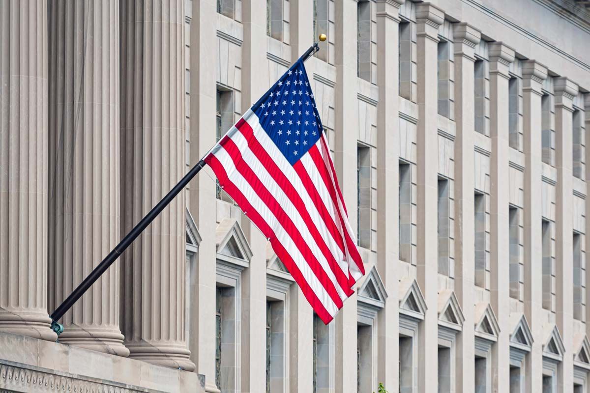 American flag waving in front of a government building with columns and windows.