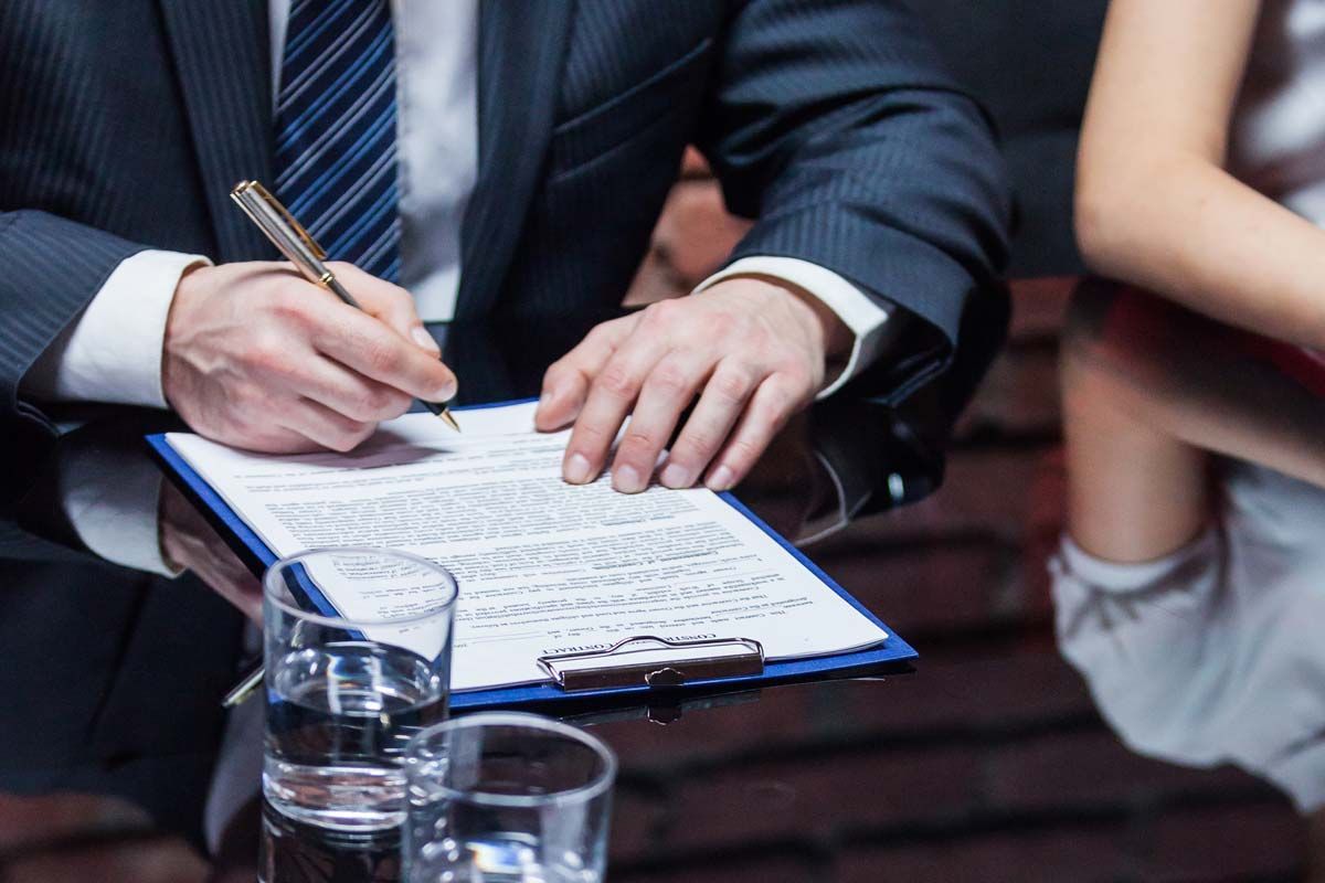 Person in suit signing document on a clipboard. Two glasses of water sit on the table nearby.
