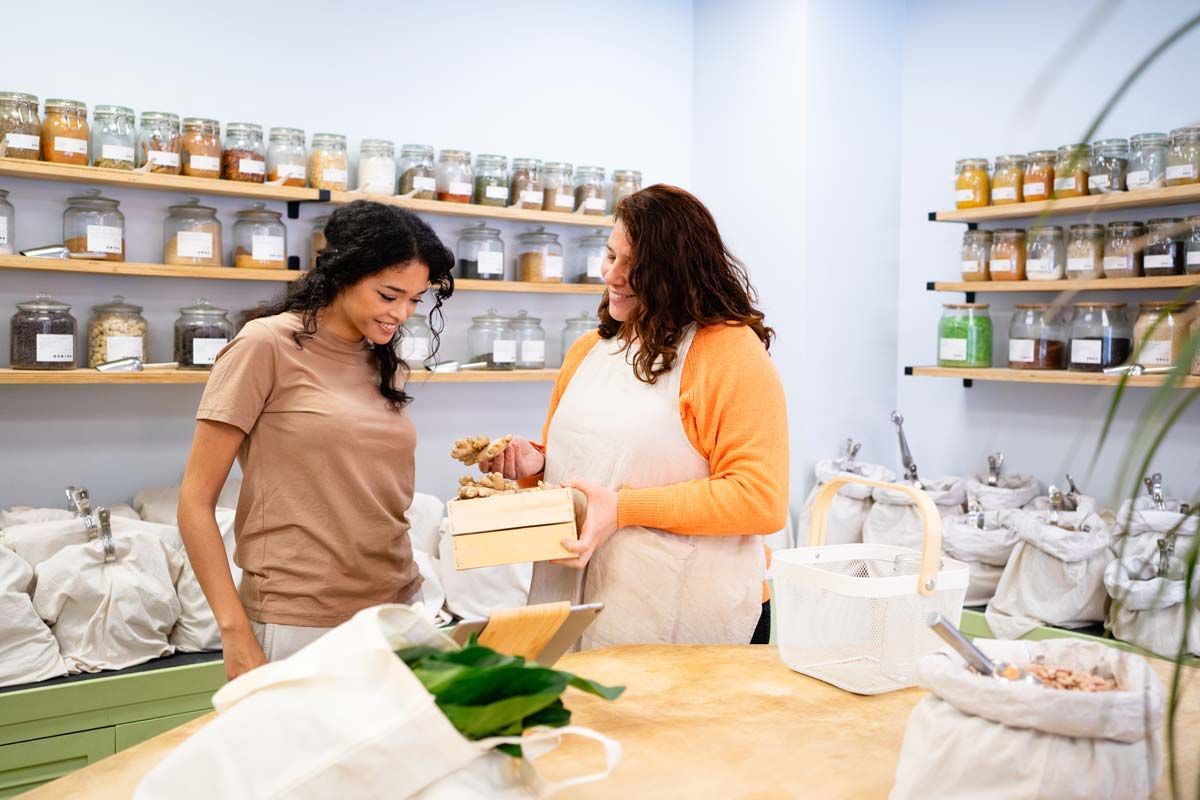 Woman shopping at a zero-waste store, interacting with a store employee behind a counter filled with food products.