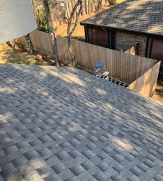 High-angle view of a gray shingled roof overlooking a backyard with a wooden fence, trees, and a neighbor's house.