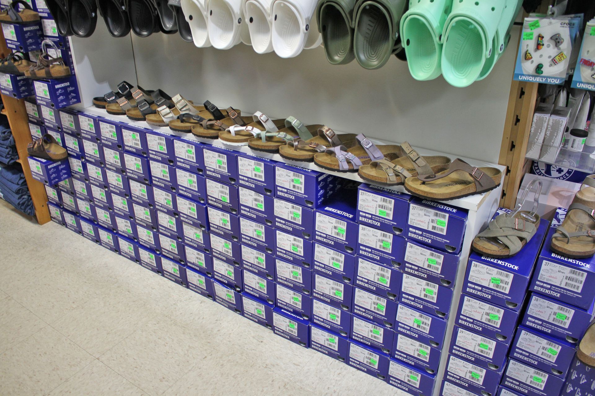 Shoes on display in a store, including sandals and Crocs on shelves. Boxes of shoes below.