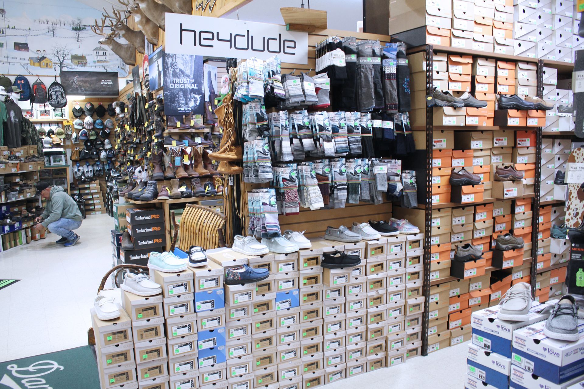 Shoe store interior with Hey Dude footwear display. Boxes stacked as shelves, various shoes on display. Person browses.