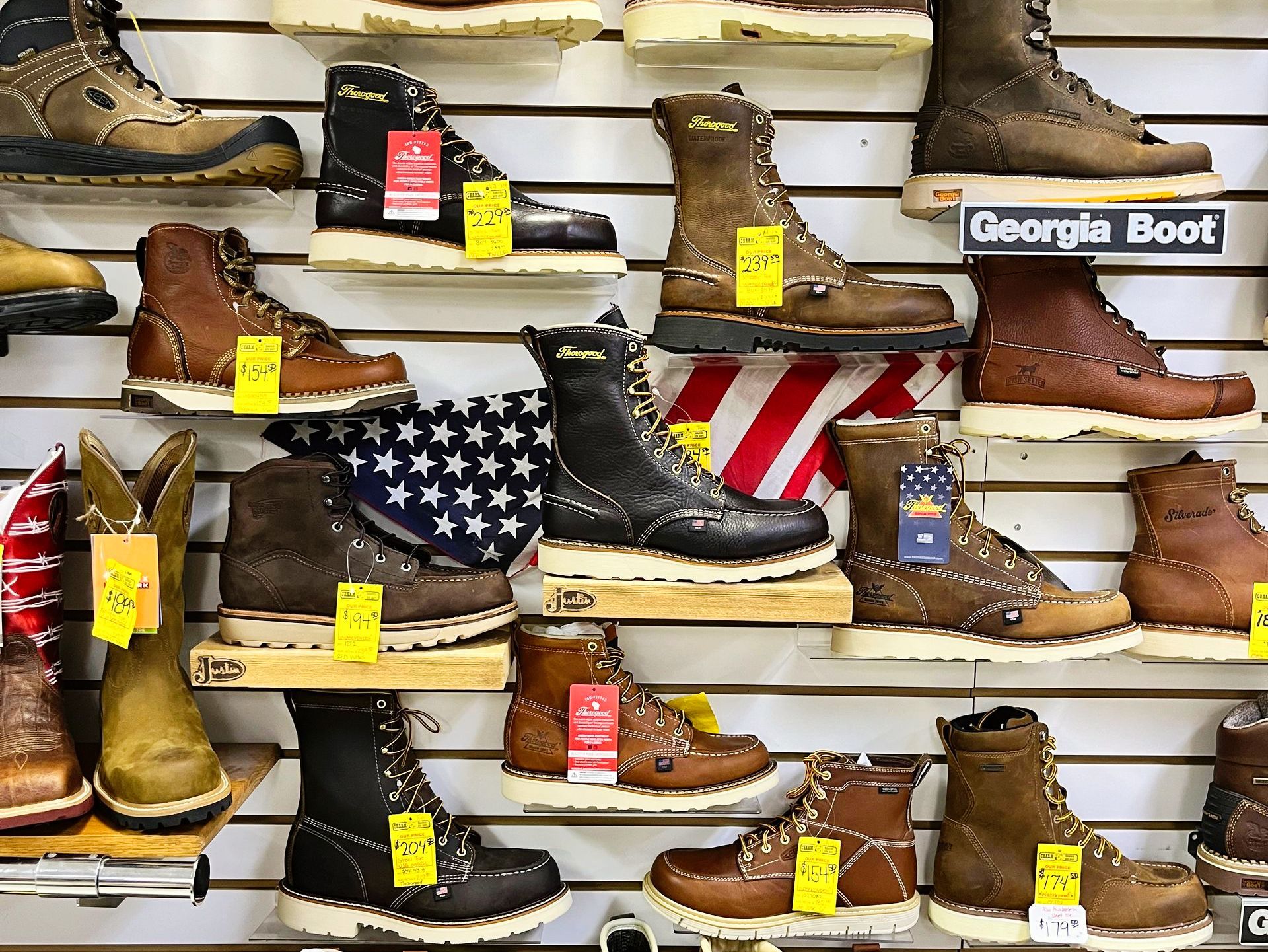 Boots displayed on shelves in a store. Various styles and colors with price tags. American flag visible.
