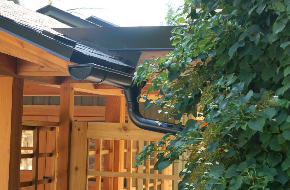 Black rain gutter on a wooden building with a dark roof, foliage in the background.