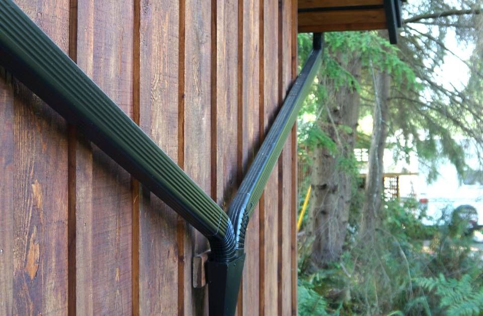 Black metal rain gutter against brown wooden siding, angled towards the right. Green trees in background.