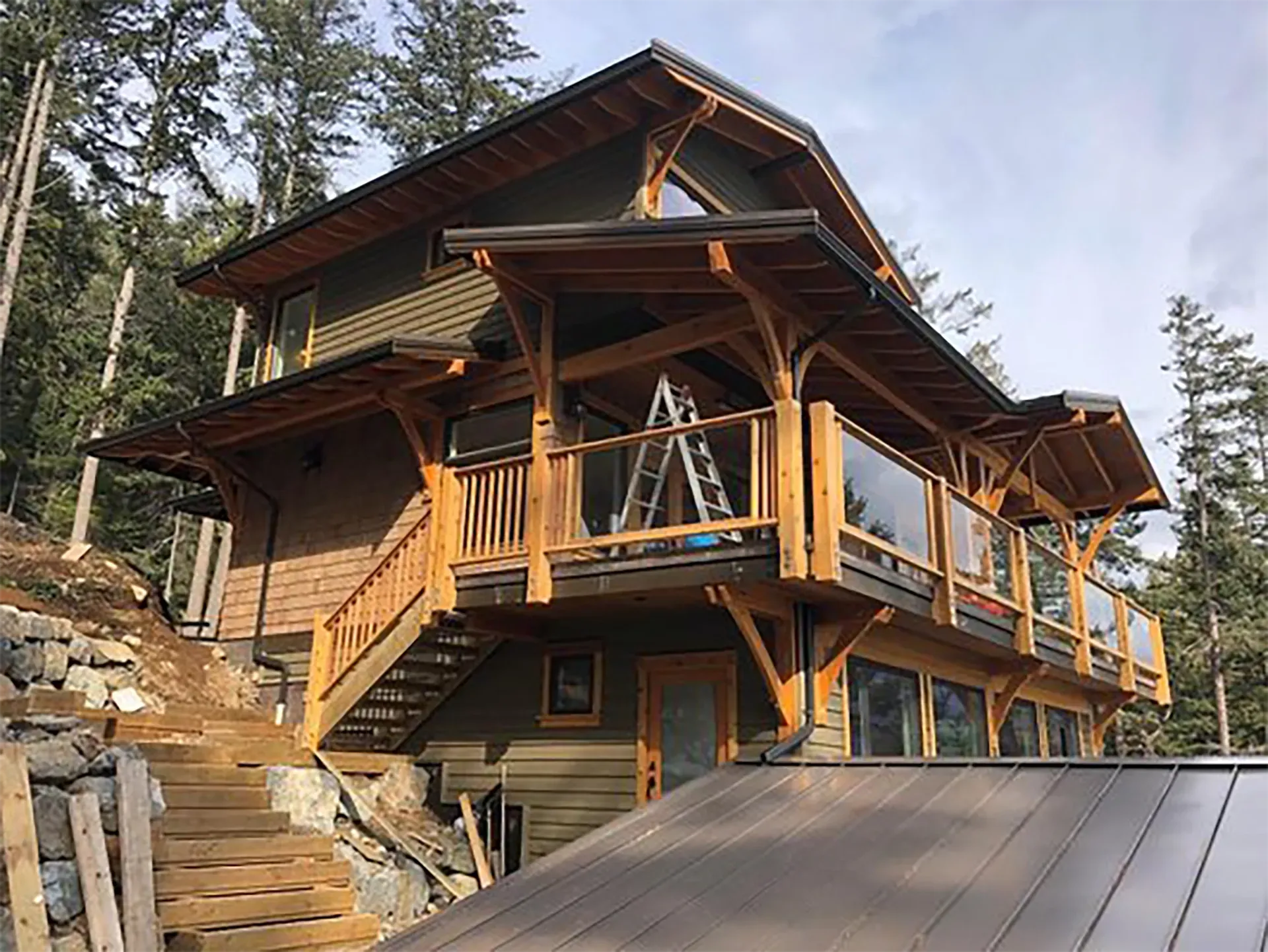 Two-story wooden house with a deck, glass railings, and a stairway built into a hillside.