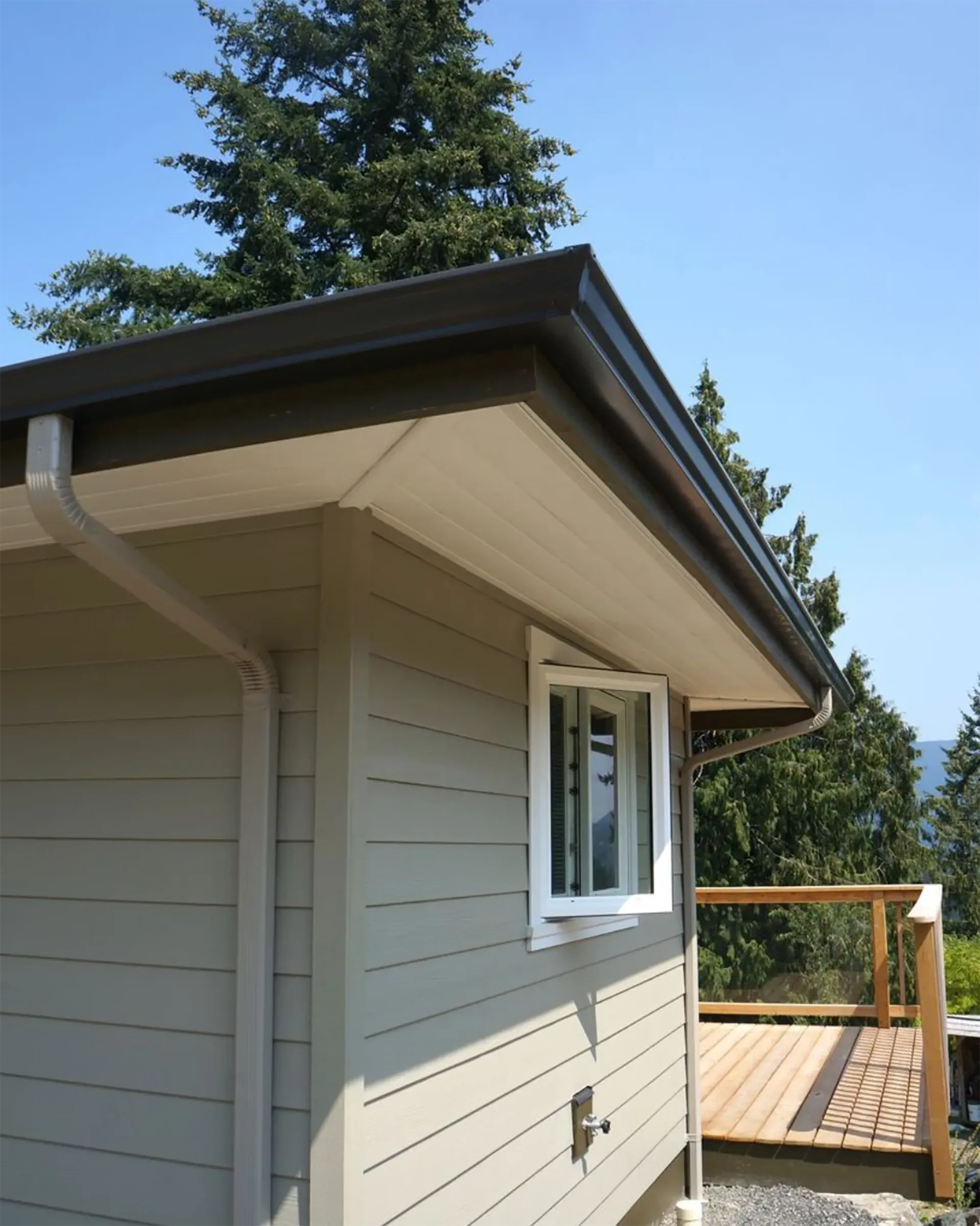 Beige house exterior with brown gutters and a white-framed window, near a wooden deck under a blue sky.