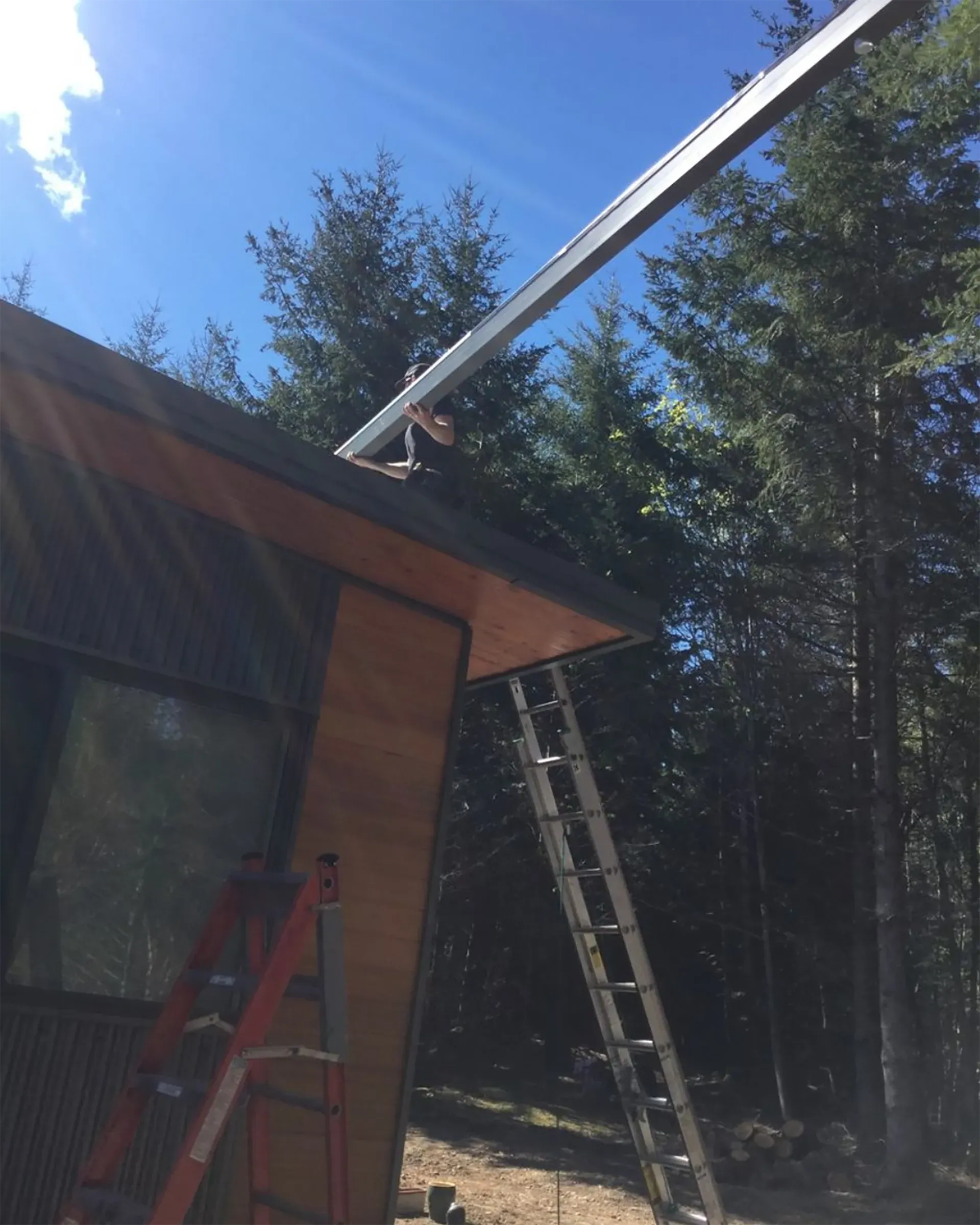A person on a ladder installs metal track for solar panels on a building's roof, outdoors. Sunny day.