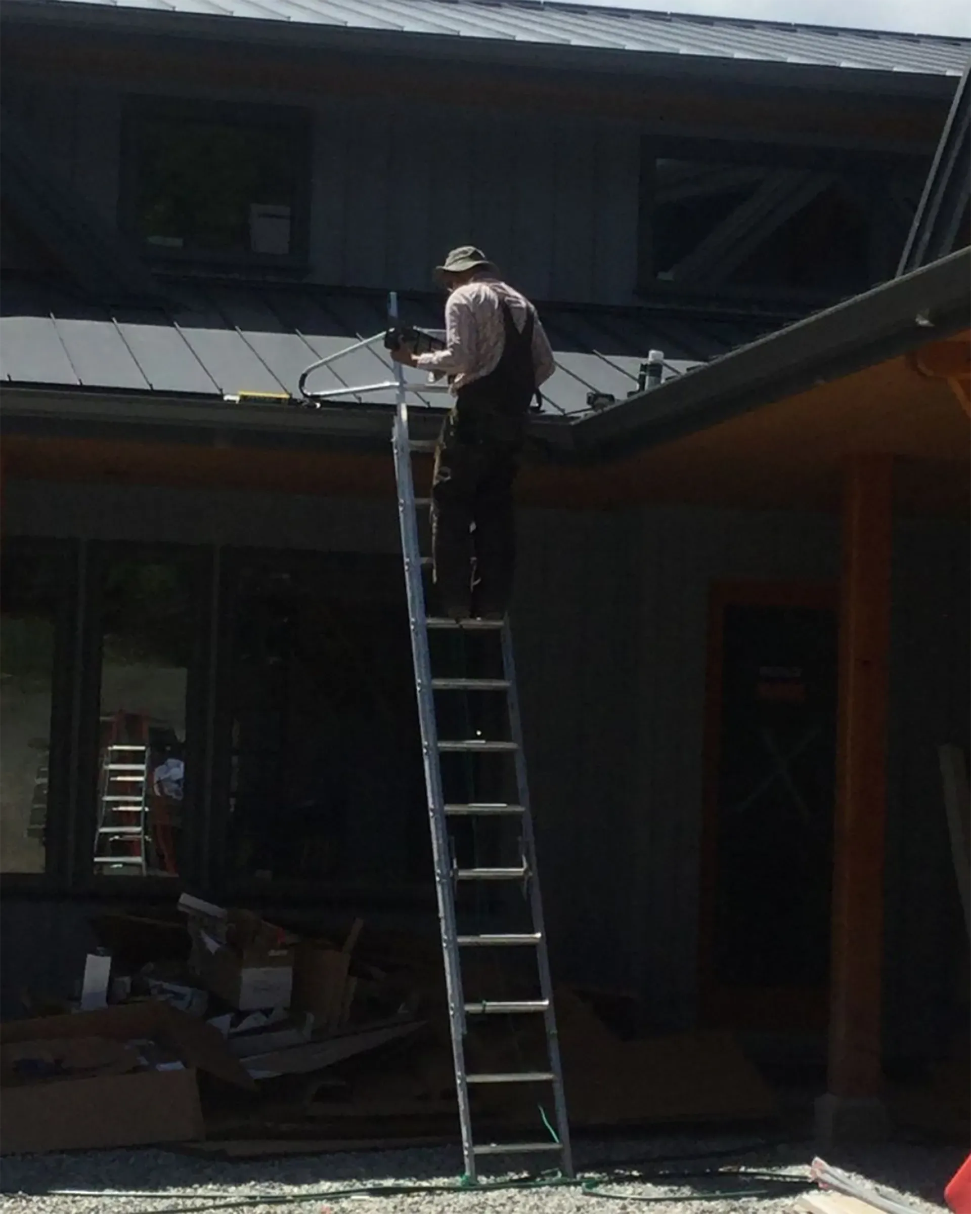 Man on a ladder cleaning a gutter on a house, under a dark roof.