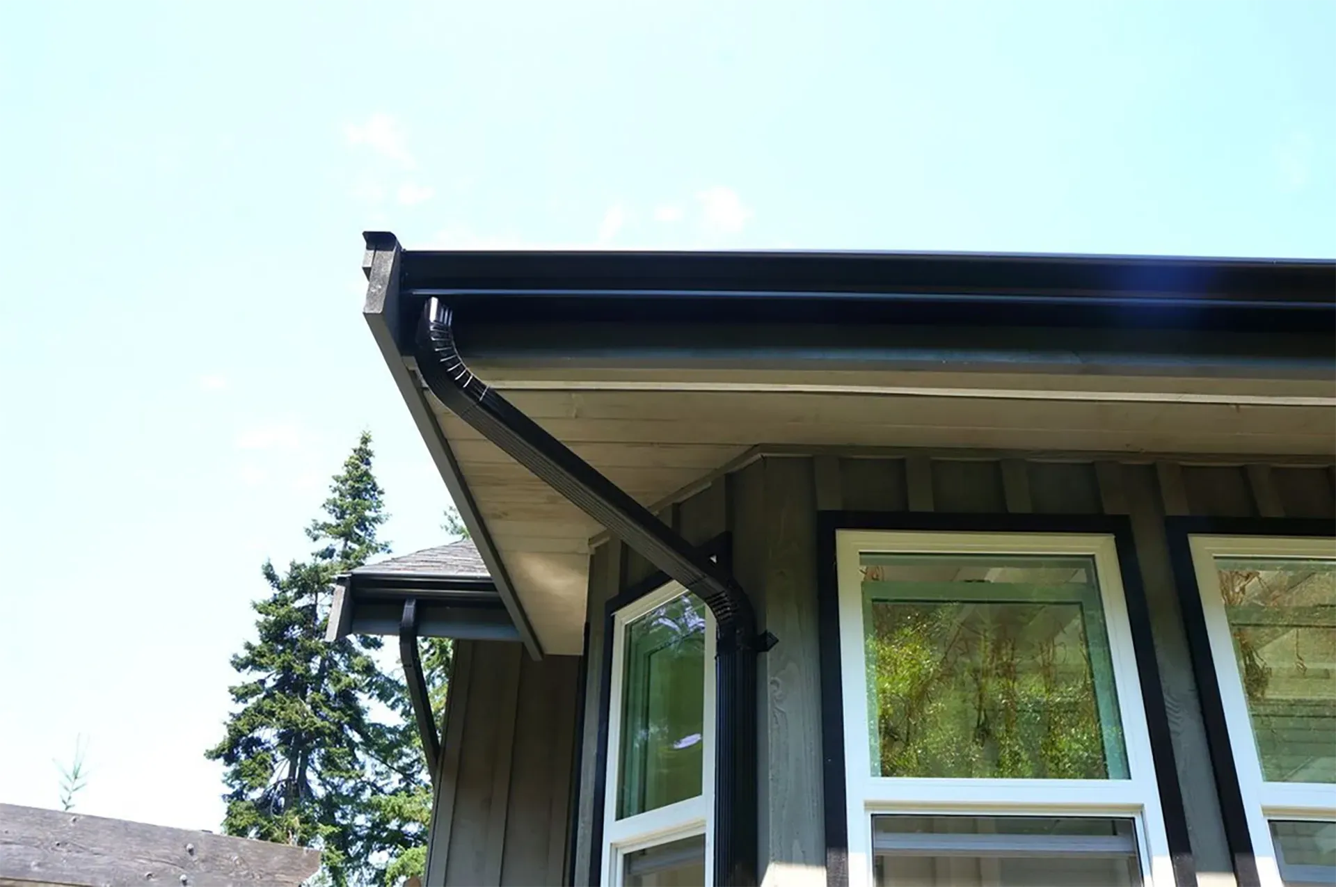 Black rain gutter on a gray building with white-framed windows, against a bright blue sky.