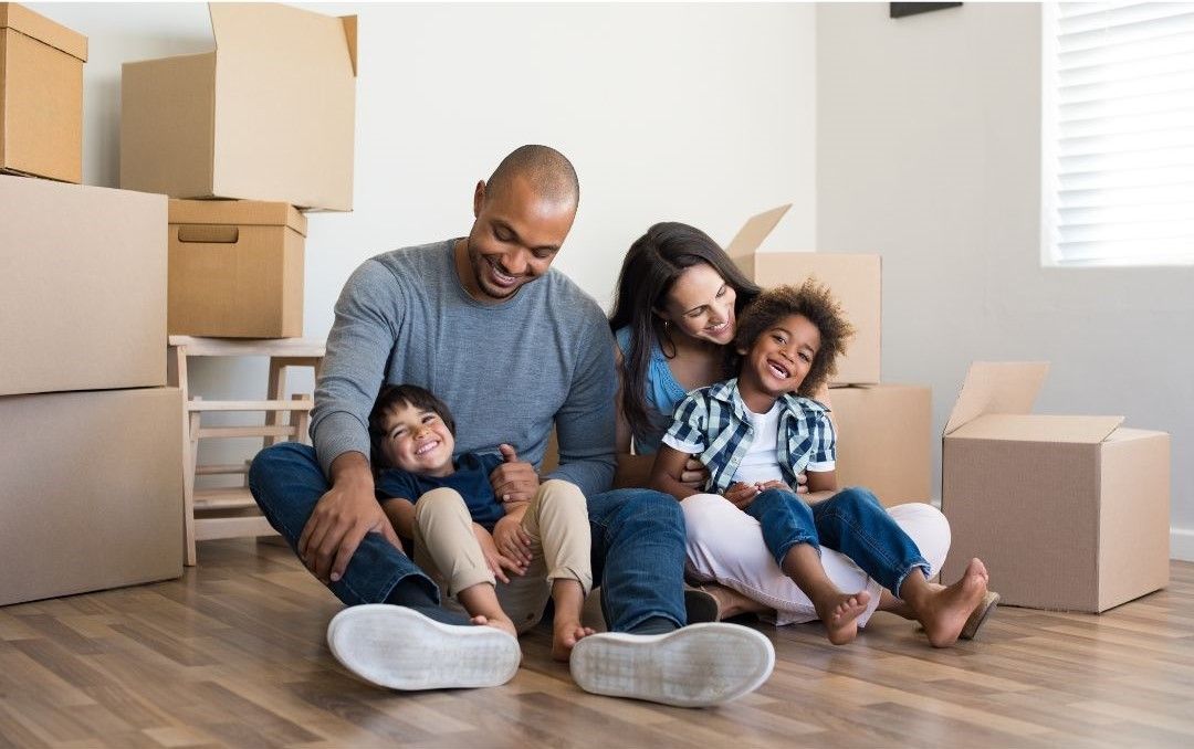 A family is sitting on the floor in a room with cardboard boxes.