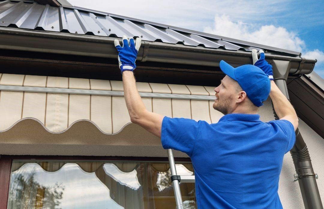 A man is standing on a ladder fixing a gutter on a roof.