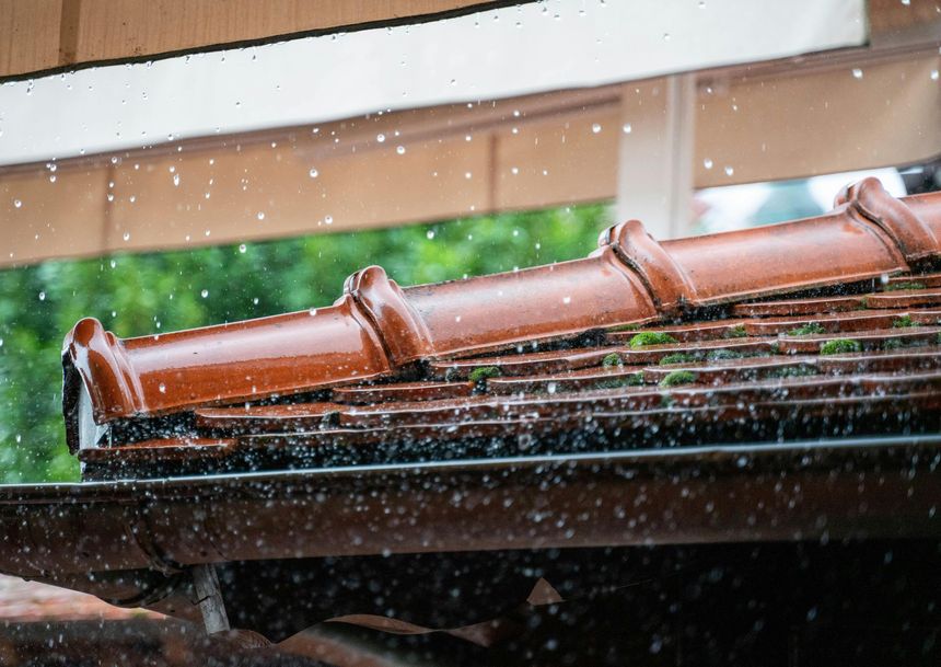 Rain falls on a brown tiled roof with a gutter, creating a mist of water droplets in front of a blurry green background.