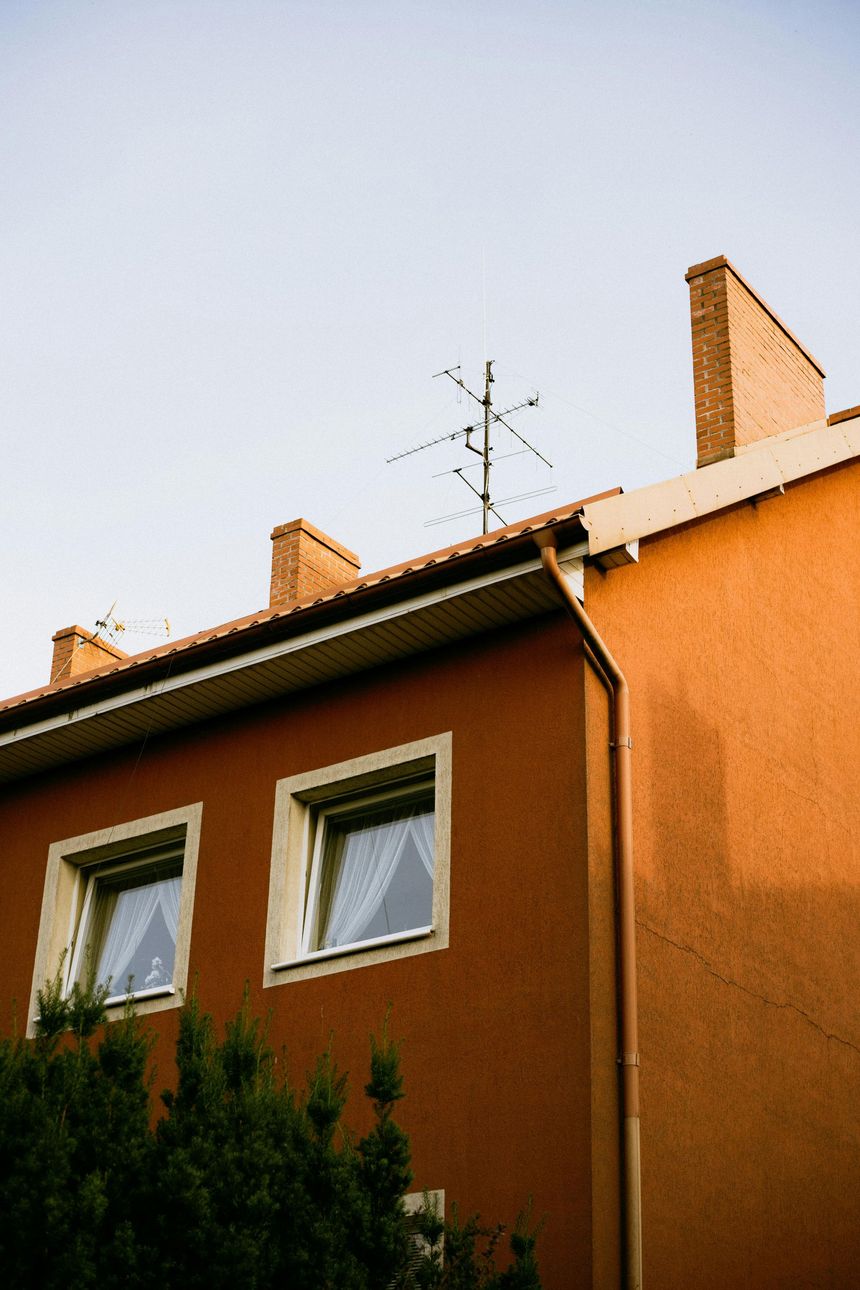 A low-angle view of a brown building exterior with two square windows, a roofline with three chimneys, and an antenna.
