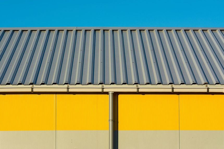 A gray corrugated metal roof sits above a wall divided horizontally into yellow and light gray panels under a blue sky.