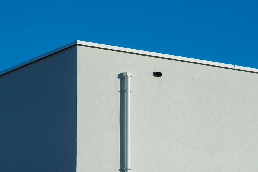 A bright white corner of a modern building against a clear blue sky, featuring a vertical pipe and a small vent.