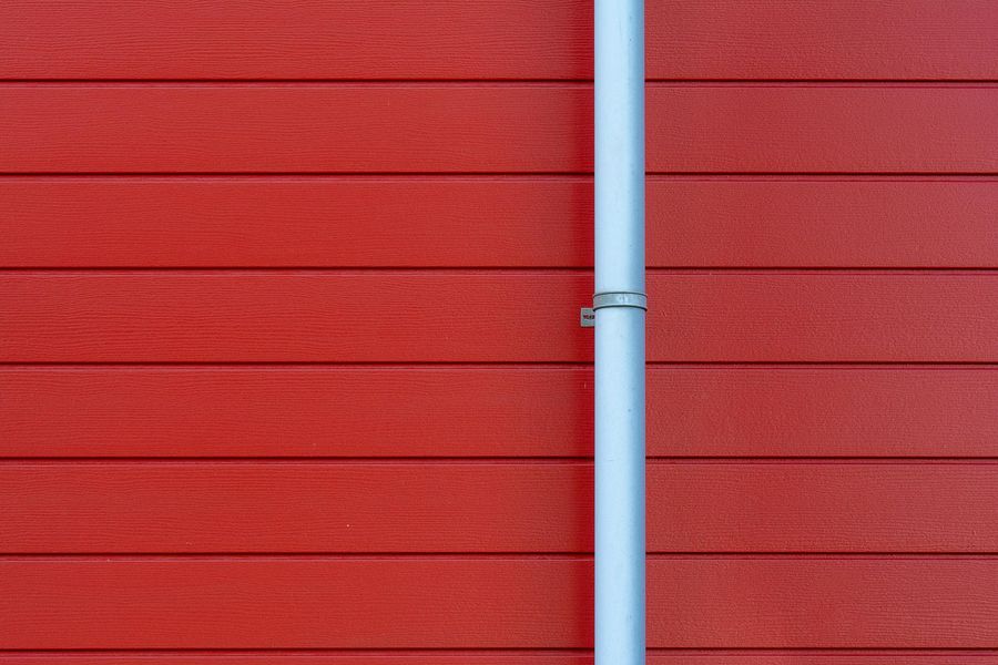 A vertical, light-colored metal pipe mounted against a wall of horizontal, red textured siding.