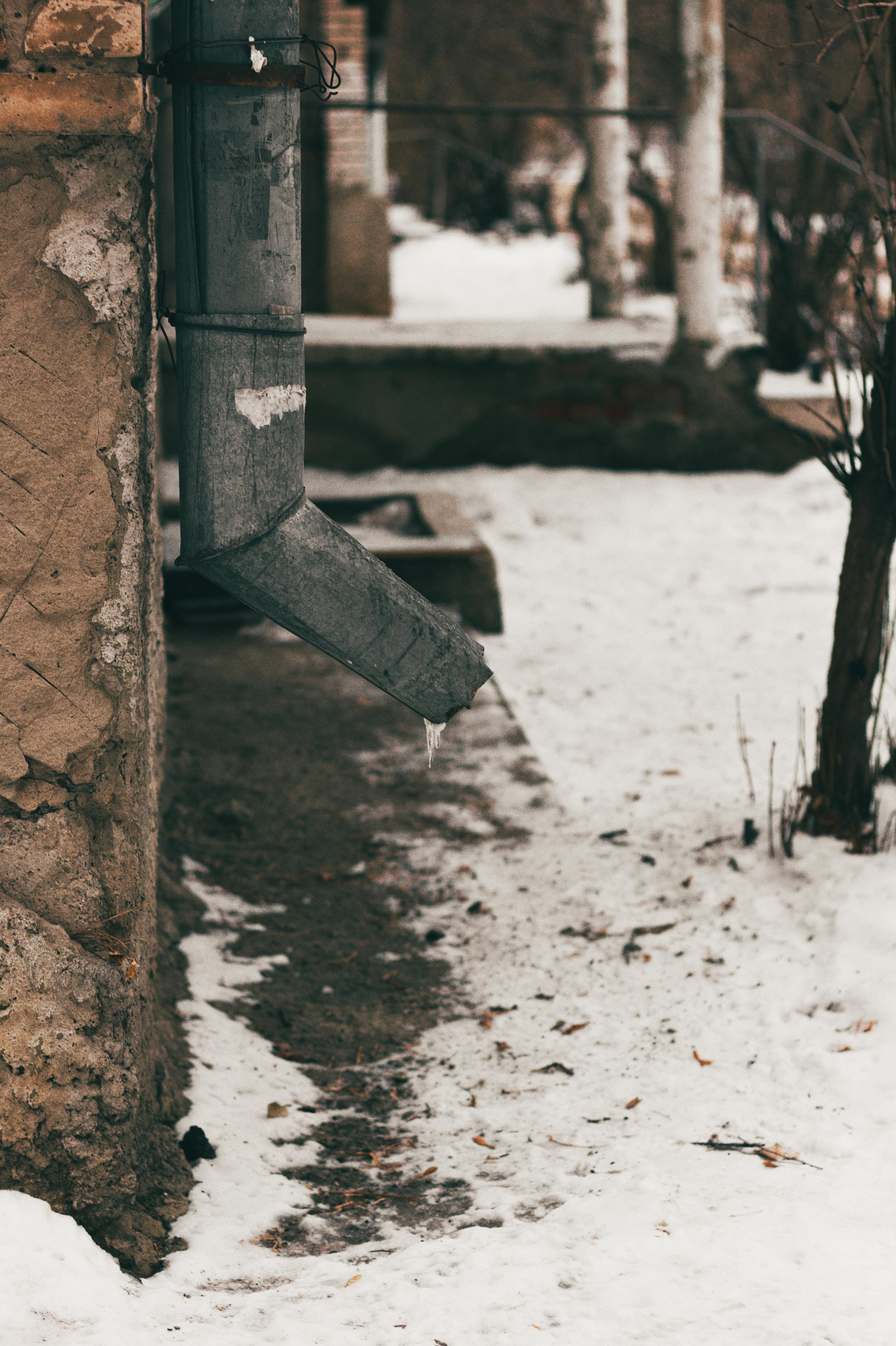 A weathered metal downspout extends from a brick building wall above a snowy ground surface.