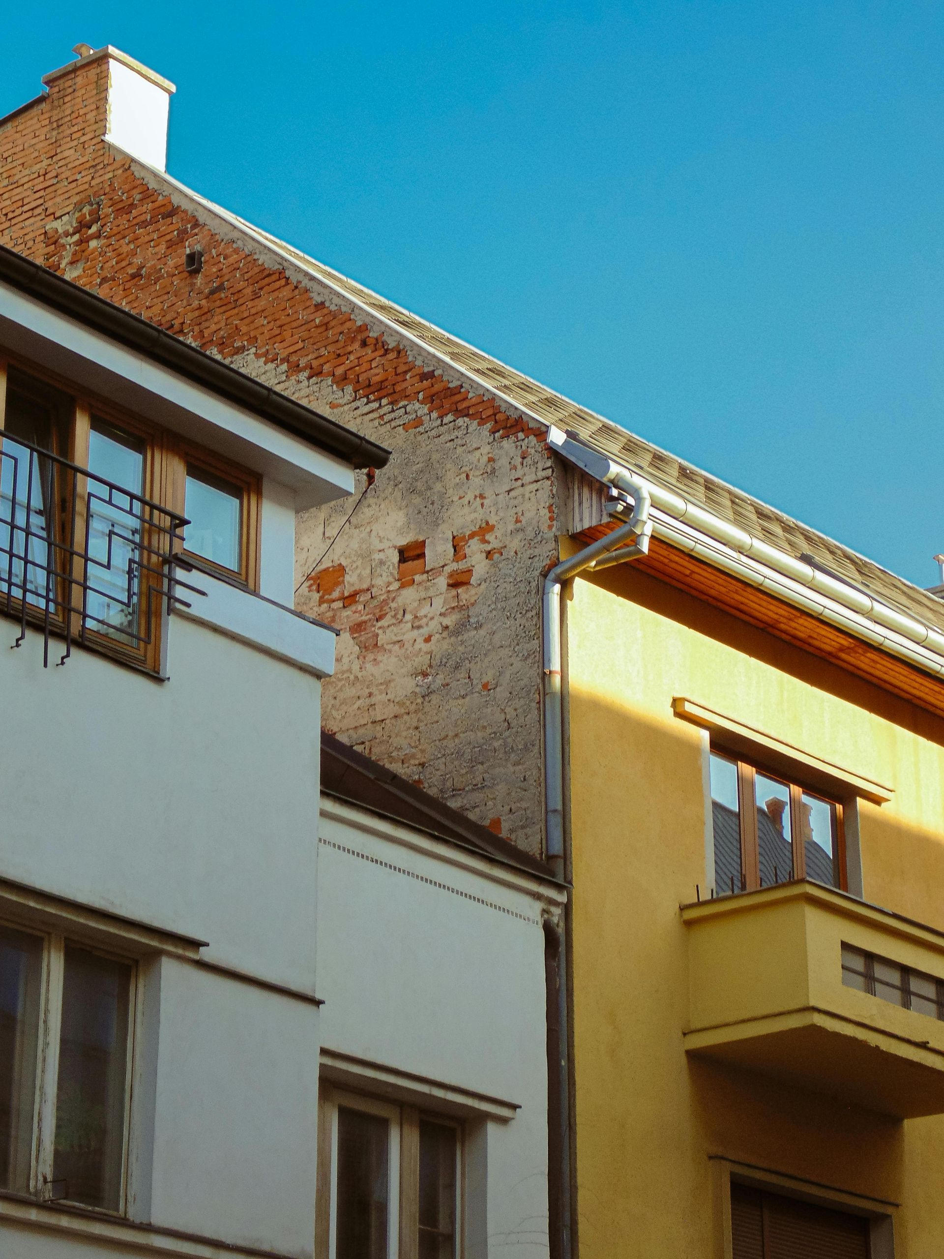 A low-angle view of two buildings with white and yellow facades, separated by a weathered brick party wall under a blue sky.