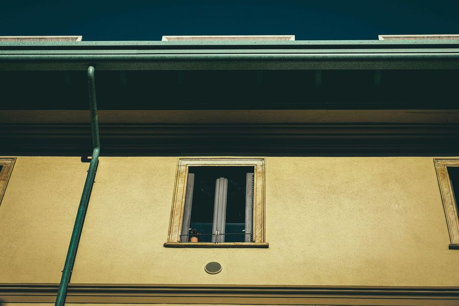 A beige building facade features a centered window with vertical curtains beneath a dark roofline and green downspout.