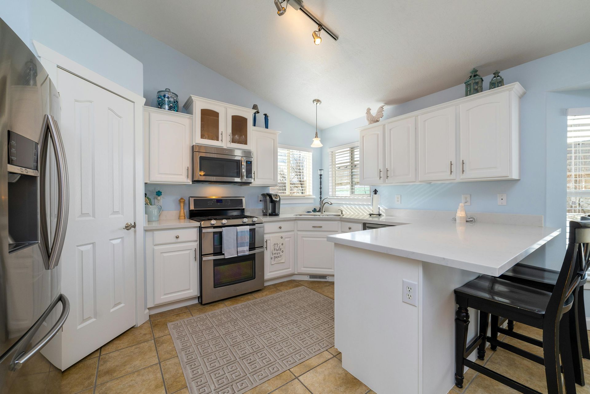 A bright kitchen features white cabinets, stainless steel appliances, a white island with black stools, and tile floors.