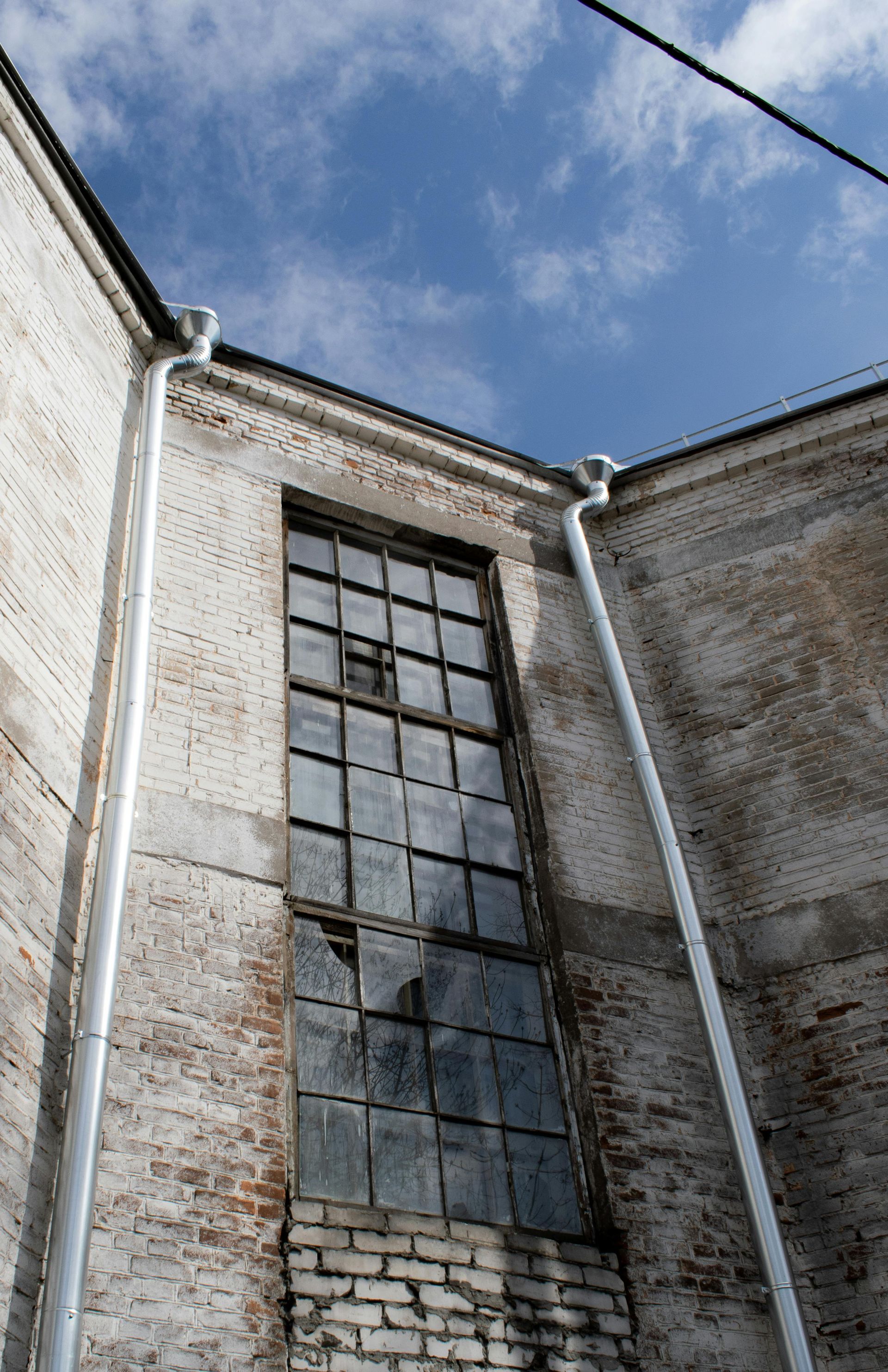 A tall, grid-paned window set into a weathered brick corner wall, flanked by two white downspouts under a blue sky.