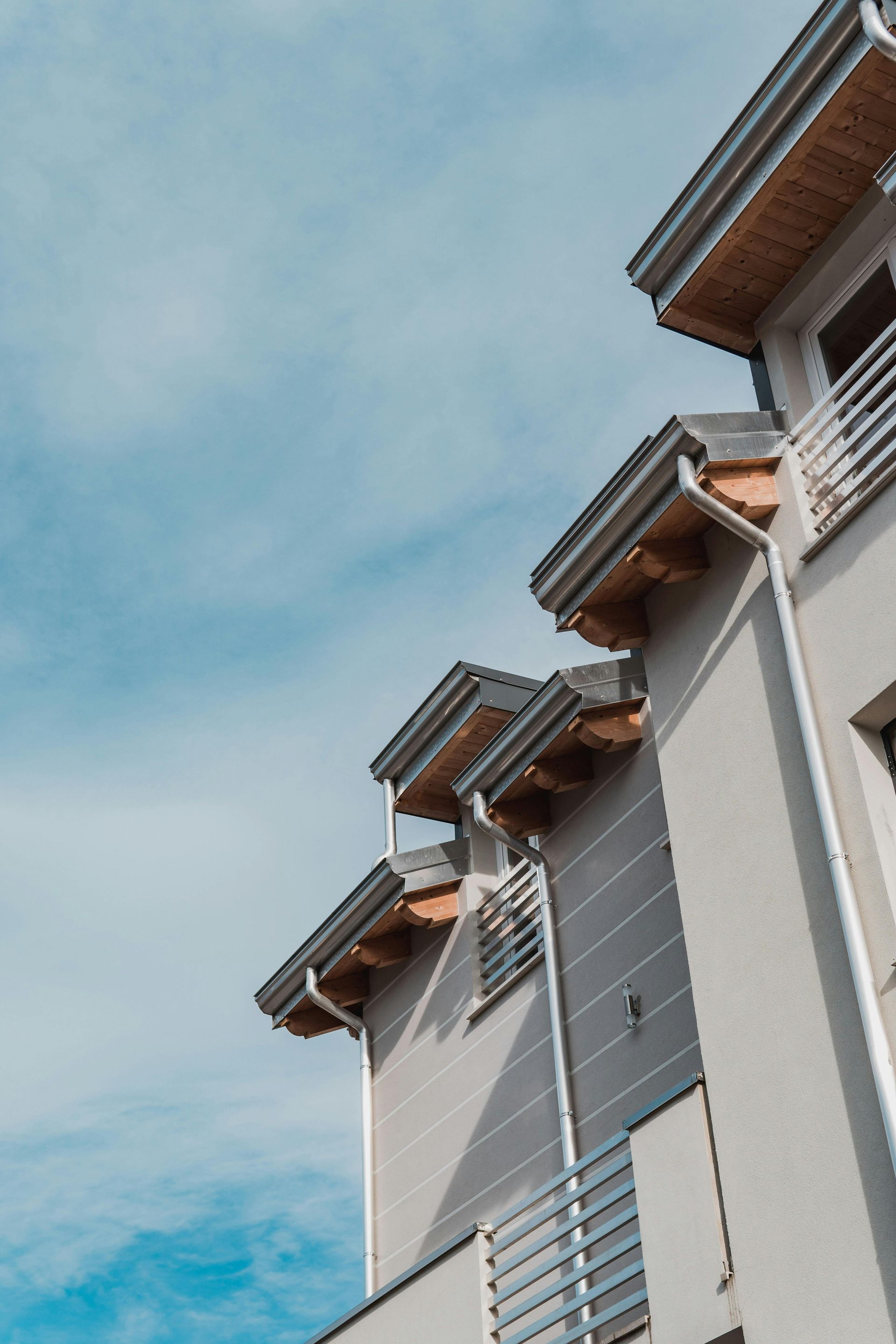 Low-angle view of the light-colored, multi-tiered roofline and balconies of a modern apartment building against a blue sky.