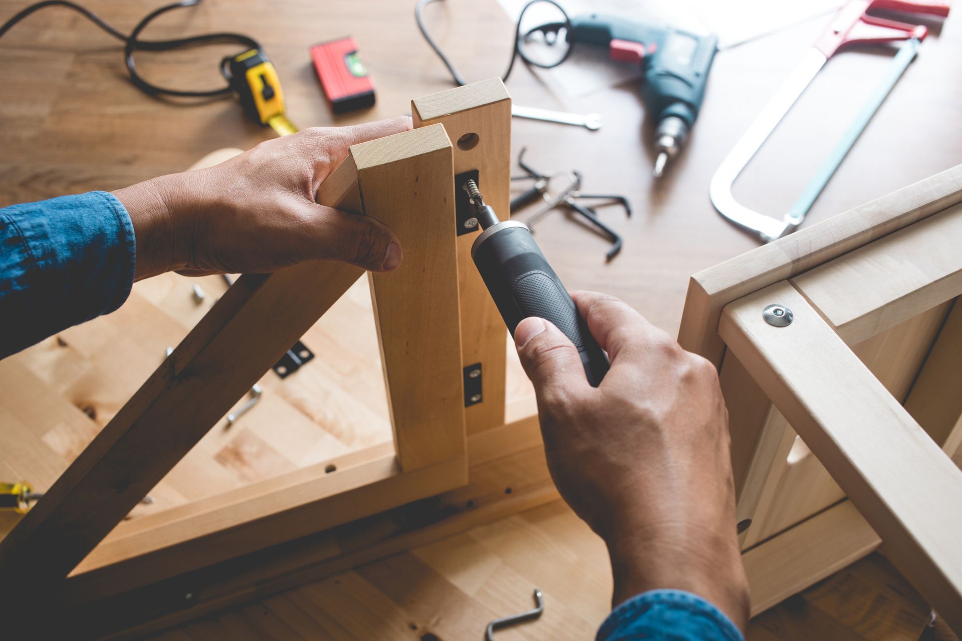 Hands using a power drill to assemble wooden furniture on a wood table, with tools scattered around.