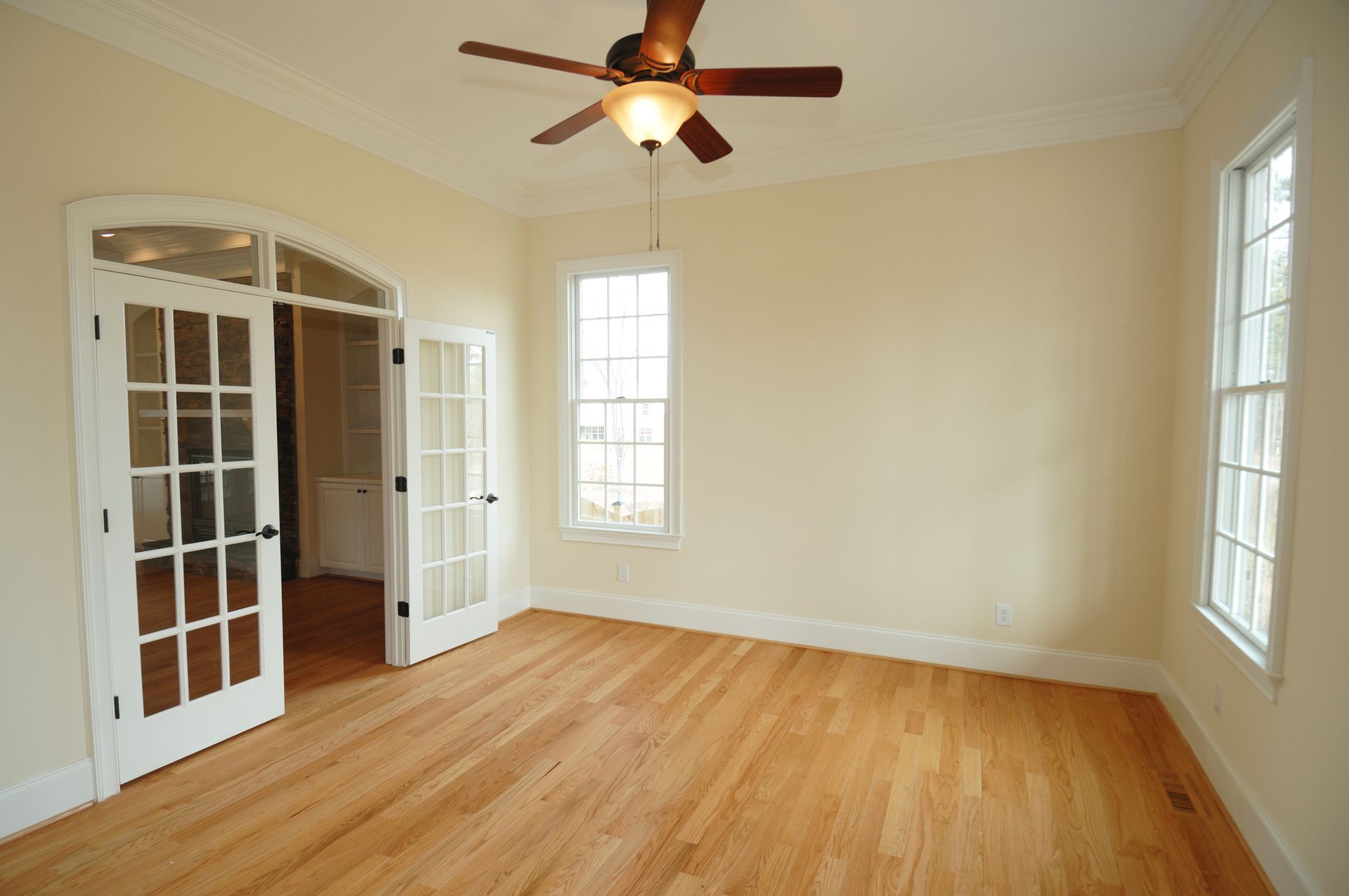 Empty room with hardwood floors, light walls, and a ceiling fan; glass doors and windows.