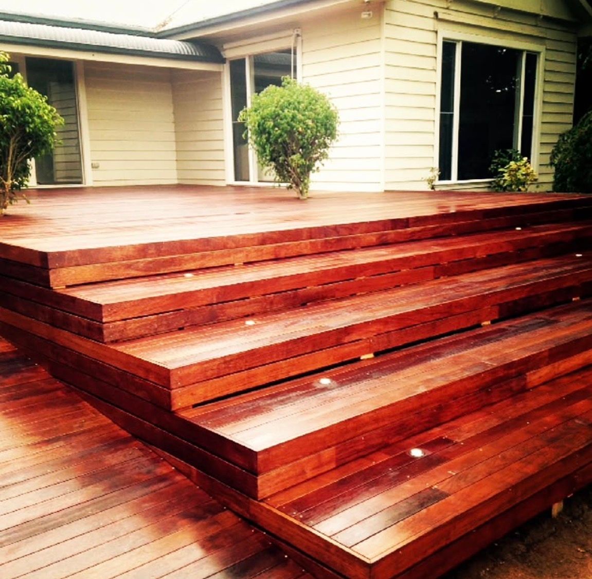 Wooden deck with steps leading to a house. Dark wood with a glossy finish, greenery in the background.