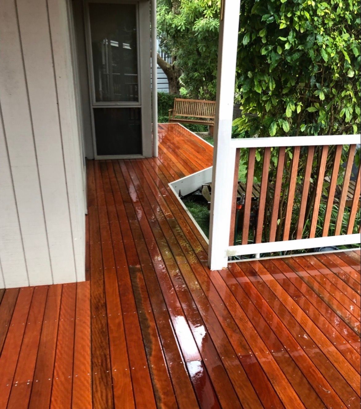 Wooden deck with wet, glossy finish; leads to doorway and green foliage.