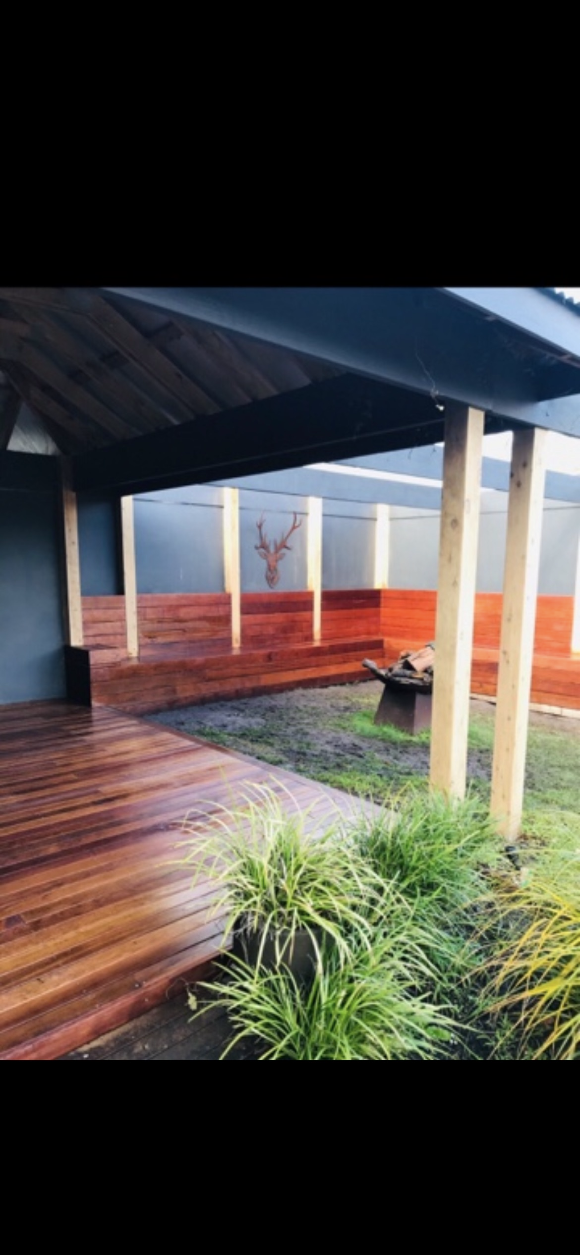 Wooden deck and benches under a black awning. Green grass and plants are in the foreground.