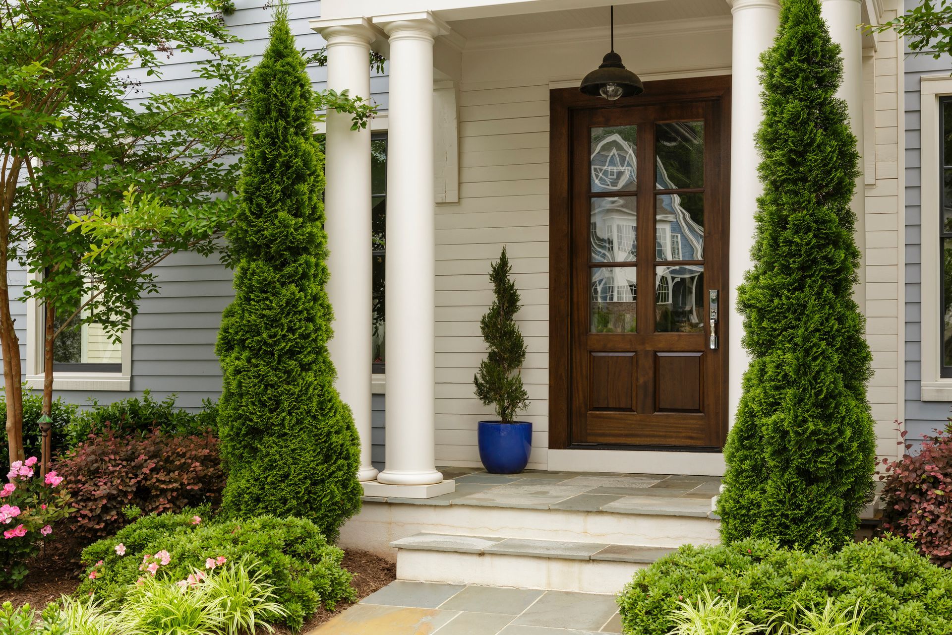 Entrance with columns, wooden door, and steps, flanked by tall green trees and shrubs.
