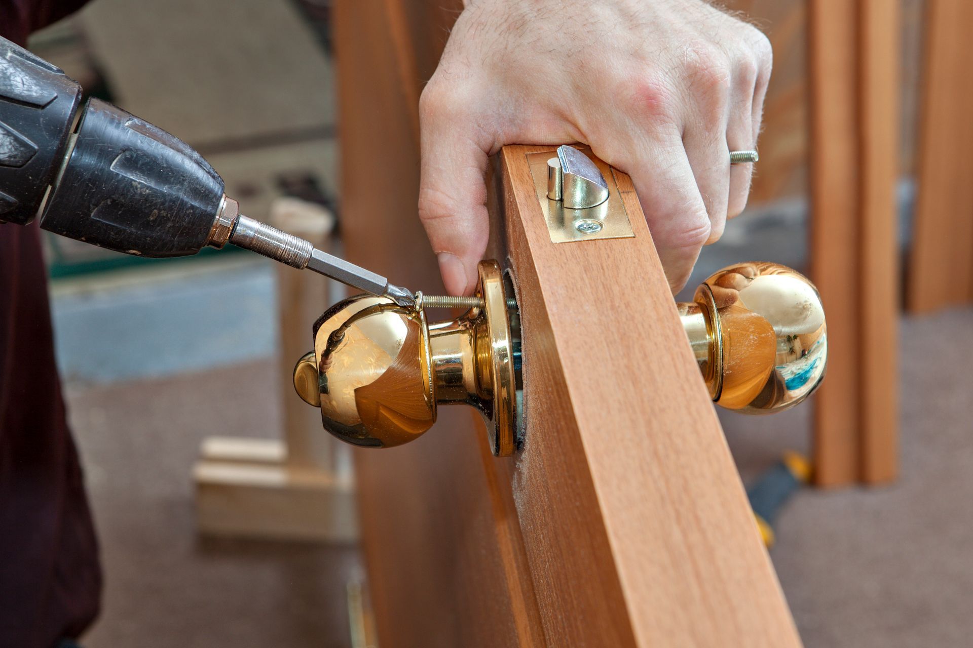 Person using a drill to install a gold doorknob on a wooden door.