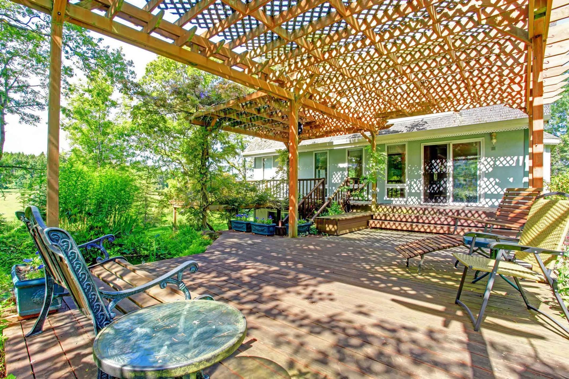 Wooden deck with lounge chairs and a table under a latticed pergola, overlooking greenery and a house.