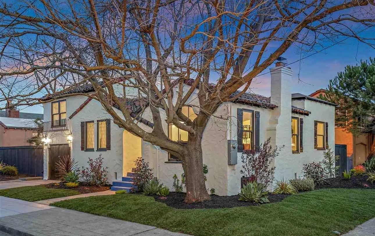 White house with a large tree in front, lit windows, and a sidewalk in a landscaped yard.