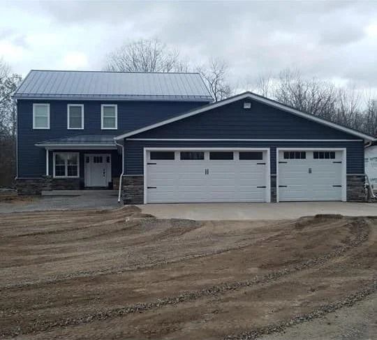 Two-story dark blue house with white trim and a three-car garage on a muddy dirt driveway under cloudy skies