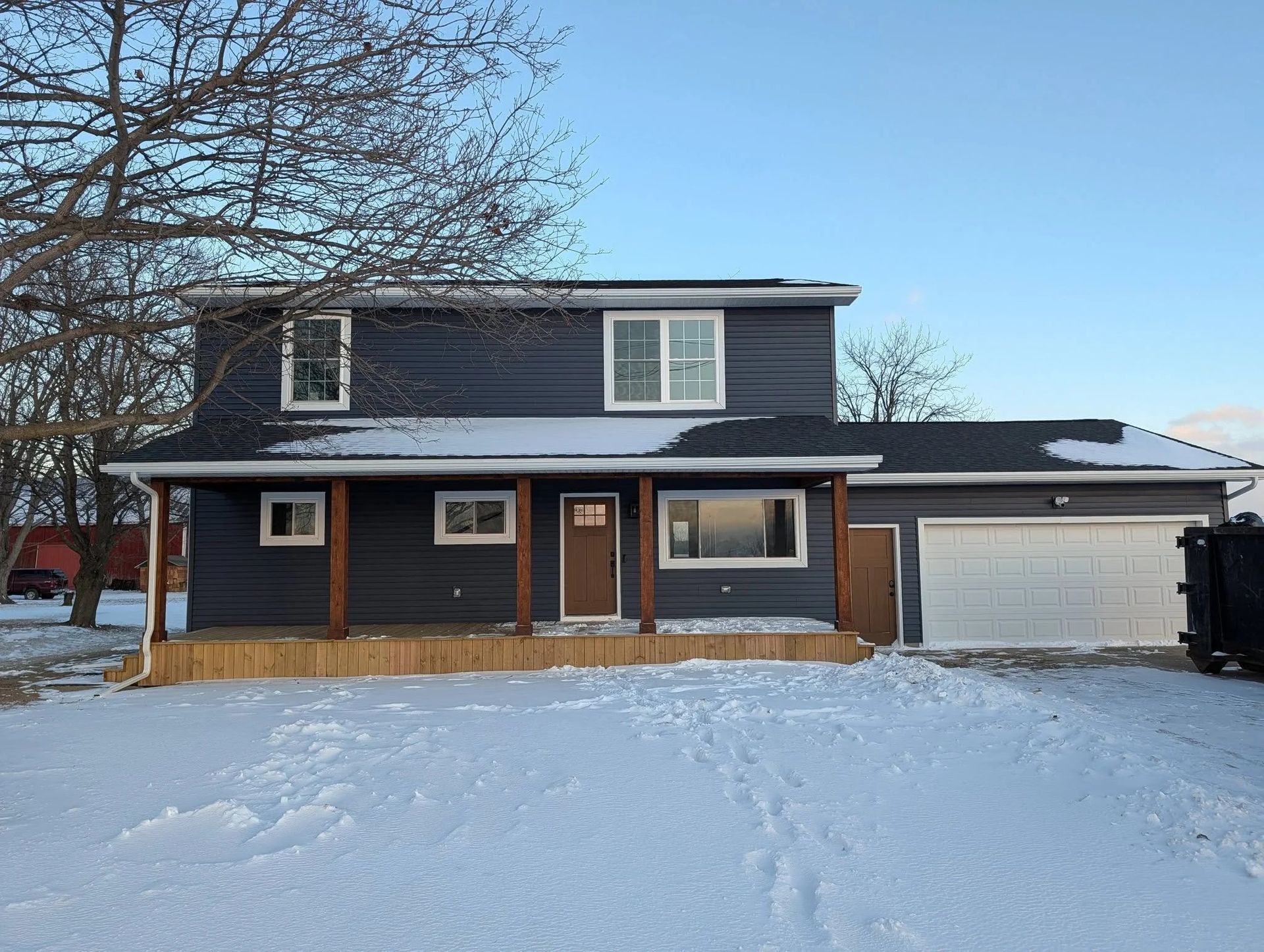 Two-story dark house with snow-covered yard, front porch, and attached garage on a winter day