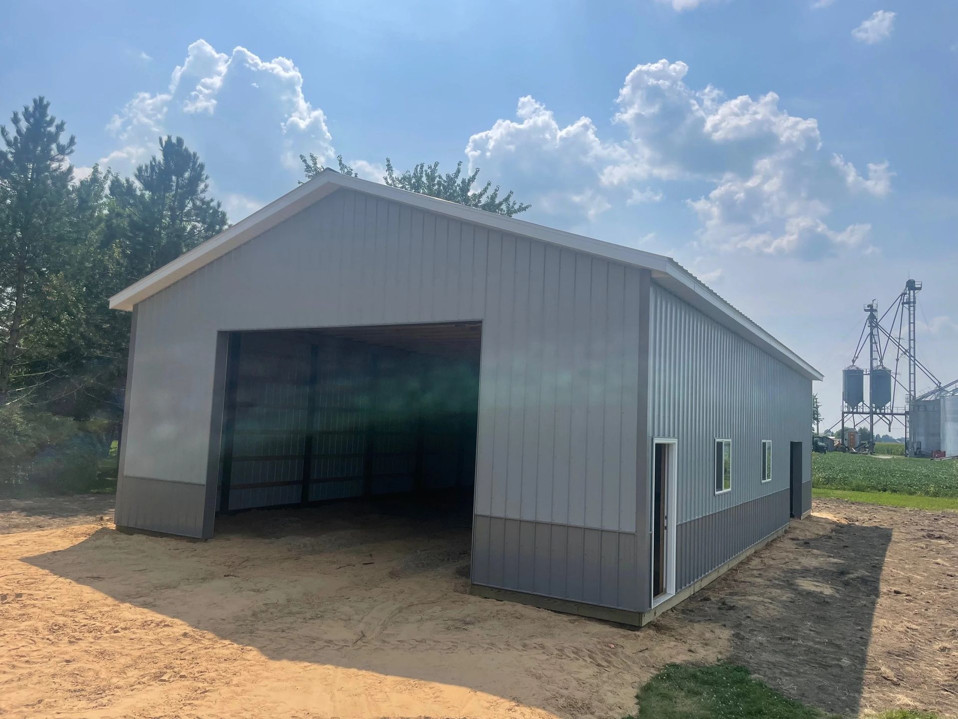 Gray metal barn-style building with large open doorway, set on a sandy lot under a blue sky.