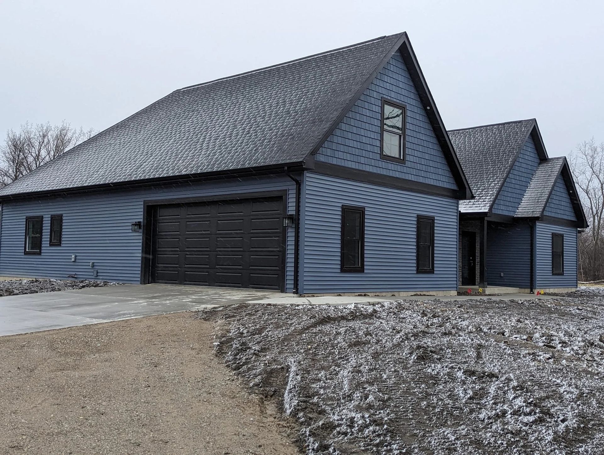 Snow-dusted blue house with a black garage and steep gabled roof on a gravel driveway