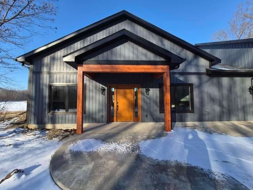 Snow-covered modern black house with wooden entryway and front steps under a clear blue sky
