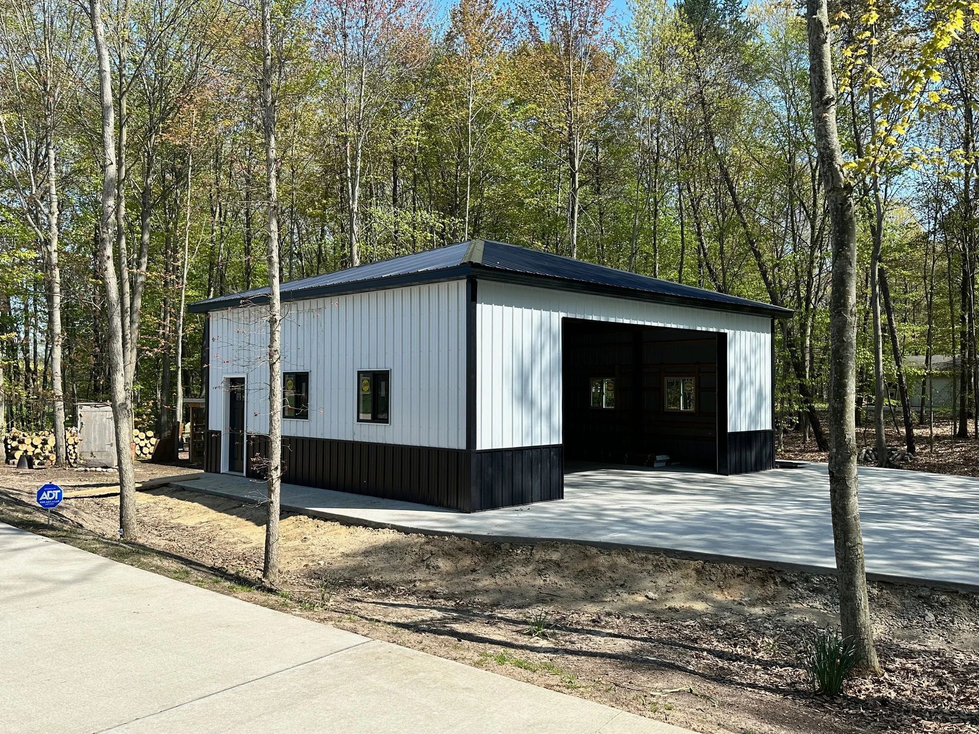 Small white metal building with a dark roof beside a wooded area and paved path