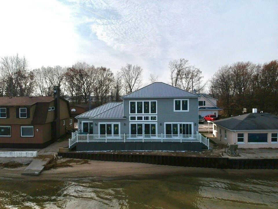 Modern gray waterfront house with large windows beside smaller homes and bare trees